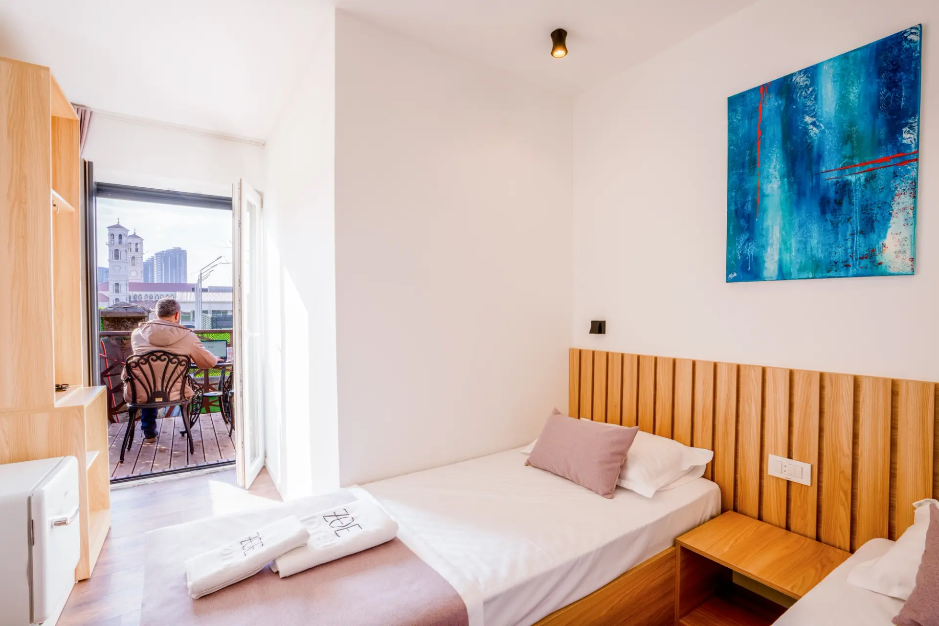 A bright ZOE Hotel room with an oak headboard, fresh linen and a blue abstract painting, opening onto a private balcony over Prishtina