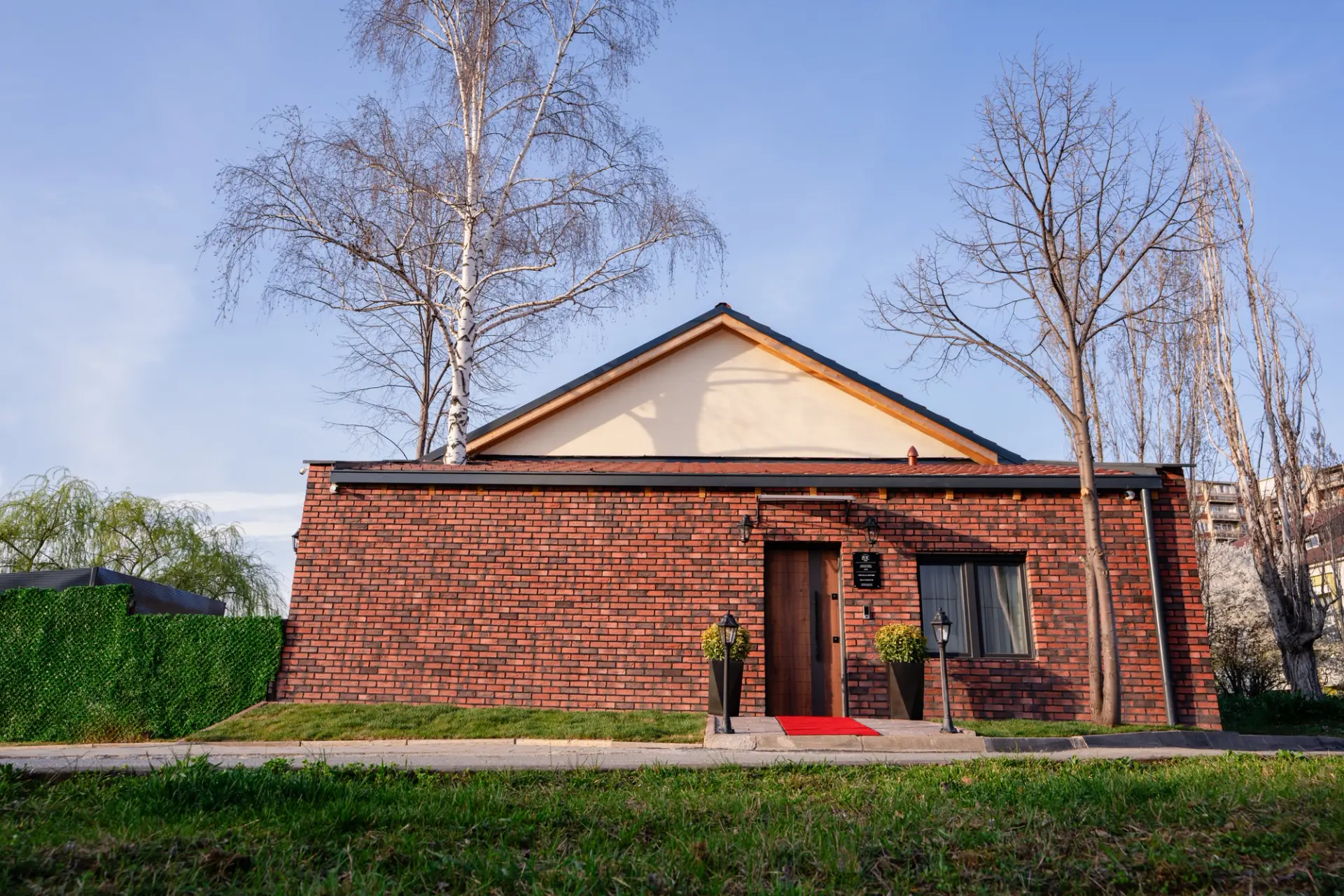 The red brick exterior of ZOE Hotel on Agim Ramadani in Prishtina, with its wooden entrance door and birch trees against a blue sky