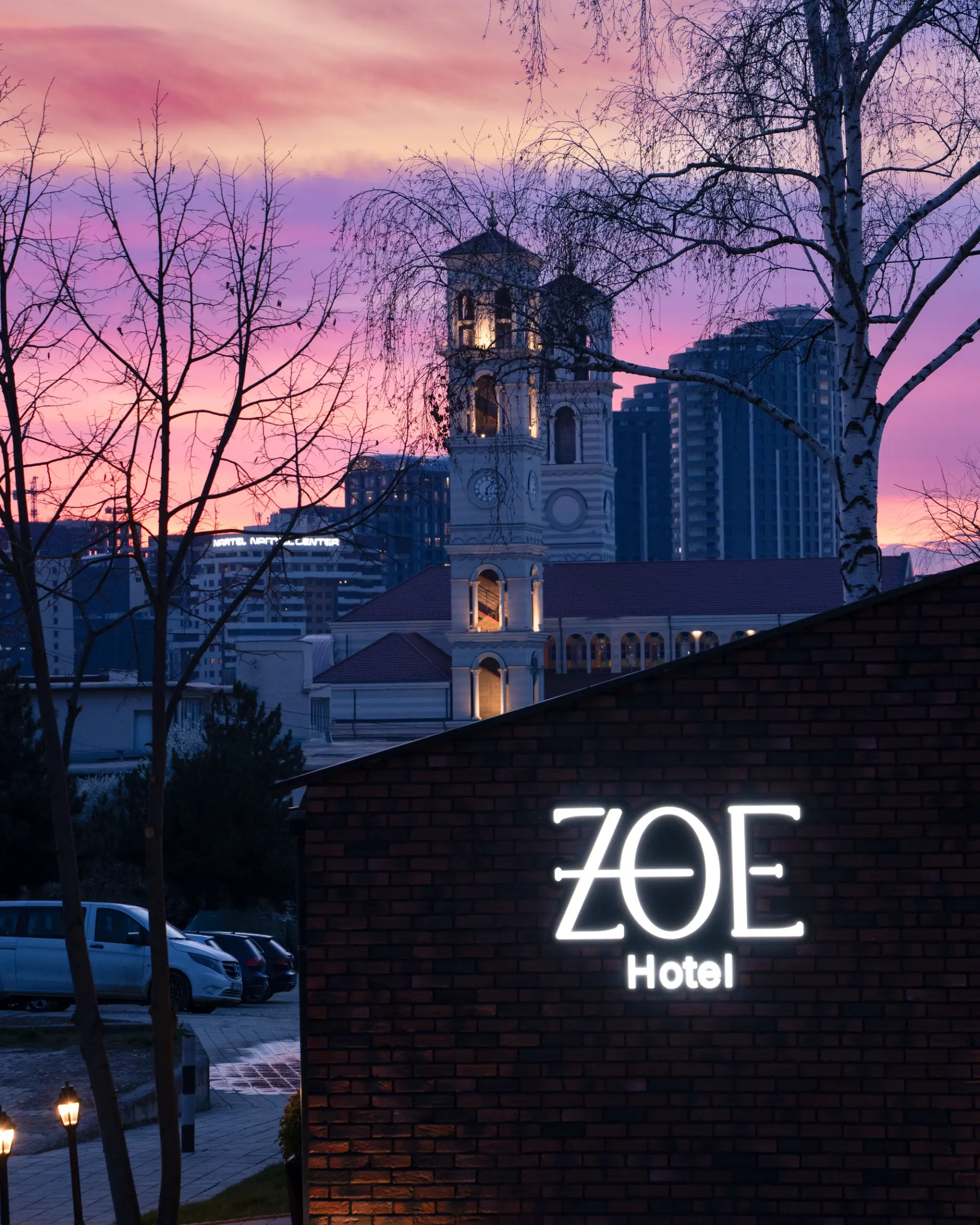 The illuminated ZOE Hotel sign on its brick wall at sunset, with the bell tower of the Cathedral of Saint Mother Teresa rising behind it in central Prishtina