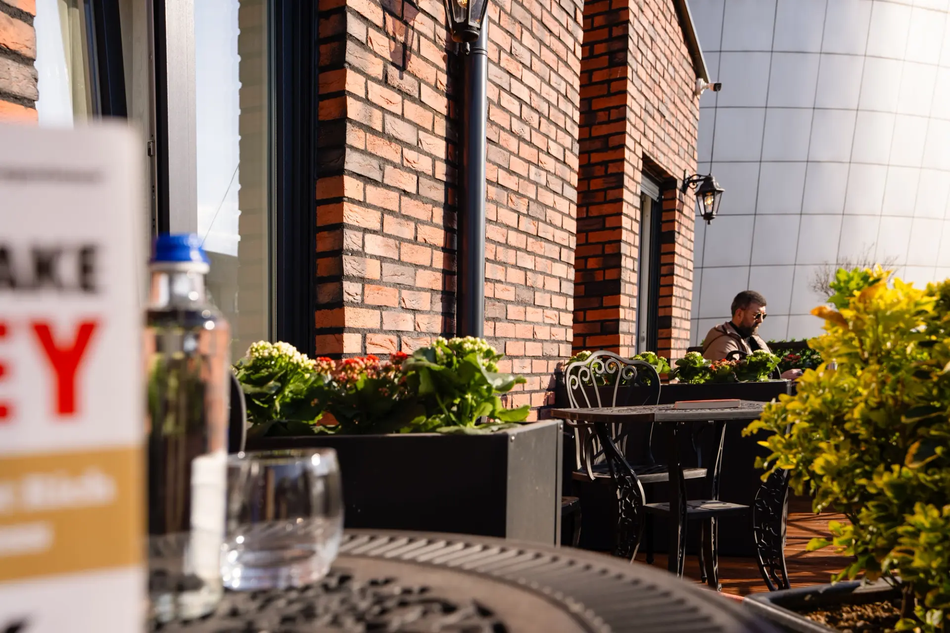 Outdoor terrace seating at ZOE Hotel Prishtina, with wrought-iron cafe tables, flower planters and a guest relaxing against the brick wall