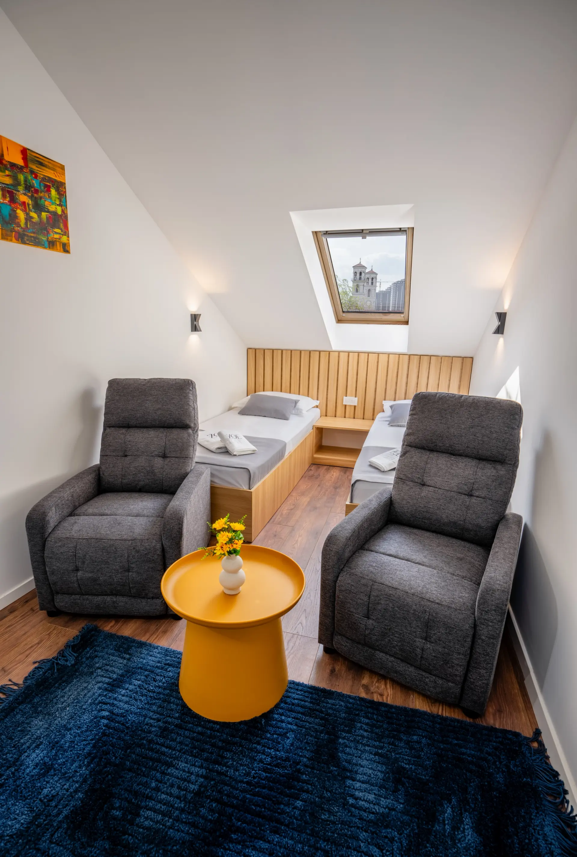 A top-floor twin room at ZOE Hotel with a sloped ceiling, two armchairs and a skylight looking out to the Prishtina cathedral tower